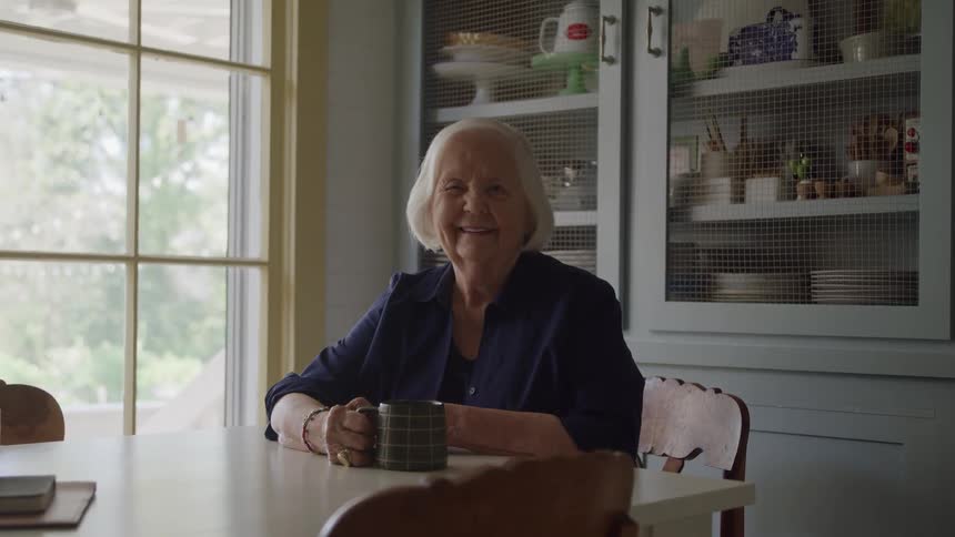 An elderly woman sitting and staring straight ahead with a smile on her face and holding a mug that is on a table