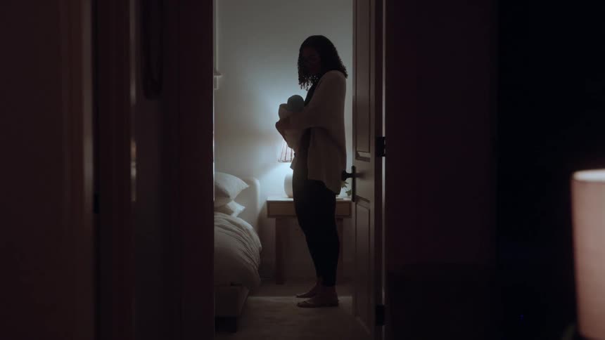 This video framed by a dark doorway shows a woman in silhouette standing in a dimly lit room gently rocking a baby wrapped in a blanket warm light from a side table which illuminates the room outlines her and the baby006