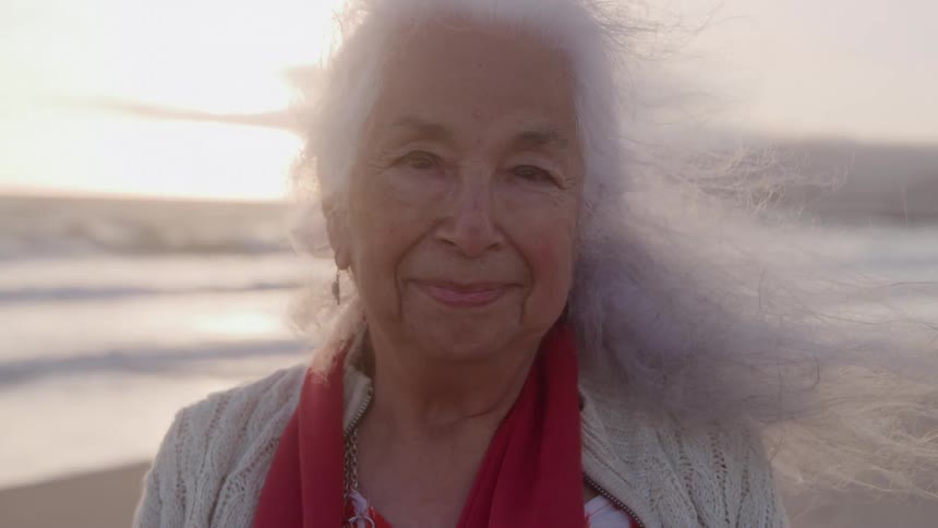 The video is a mediumcloseup shot of a smiling elderly woman with long flowing white hair blowing around her face in the wind she is standing outdoors on a beach with the ocean and a bright sunset or overcast sky visible in the background