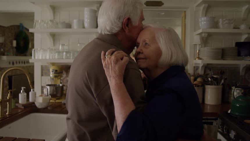 A man and a woman with white hair are hugging in a kitchen with some shelves with glasses in the background