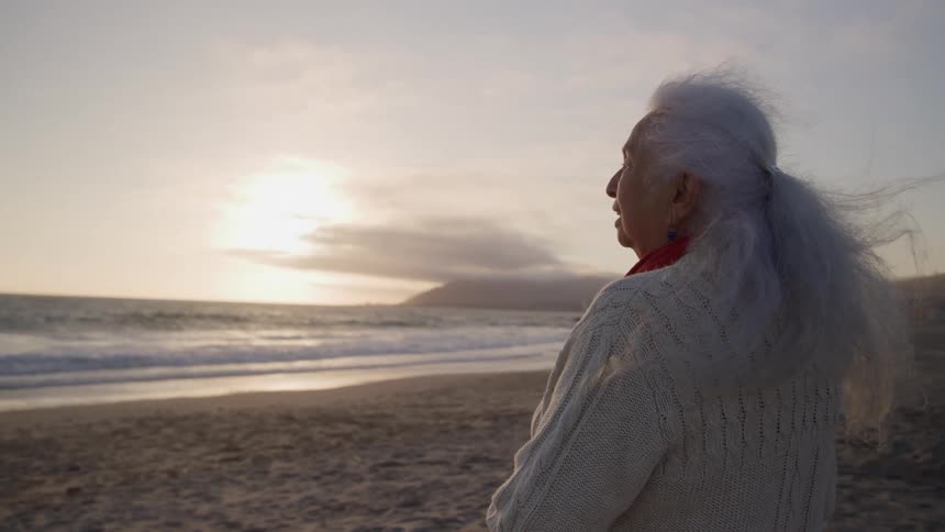 An elderly woman stands on the beach while looking out at the ocean