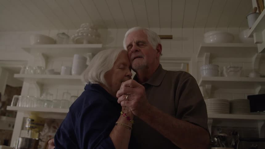 An elderly man and woman have their eyes closed as they embrace and hold hands in a kitchen