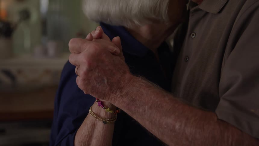 The video is a mediumcloseup shot of an elderly couple slow dancing in a dimly lit cozy indoor space the focus is primarily on their hands clasped together and the tender intimate moment of the dance