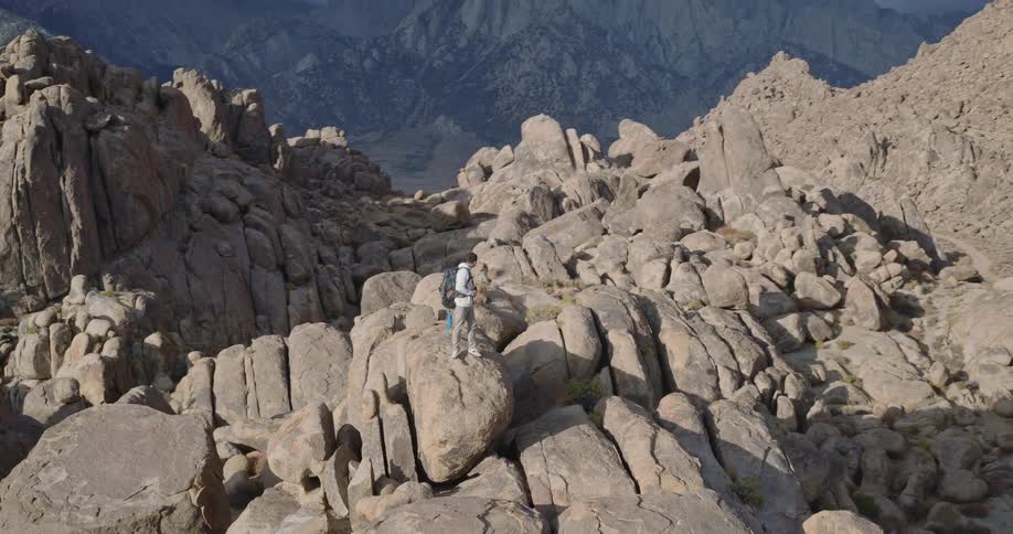 Male backpacker holds a camera while standing on a tor formation in a boulder field in mountainous terrain on an autumn day