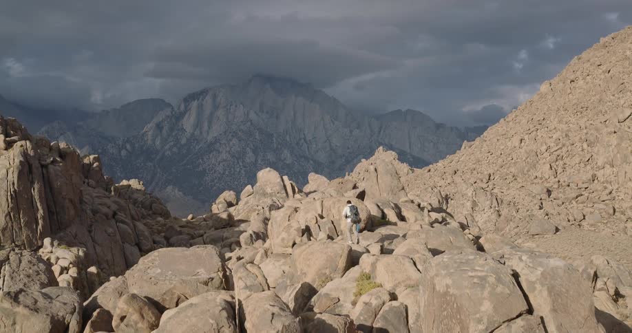 A hiker crosses rocky terrain beneath towering mountains under bright sun