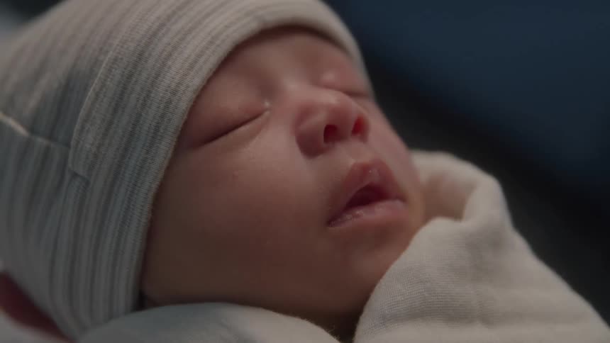A closeup shot shows a babys face wearing a lightcolored cap while sleeping with eyes closed and the lower face resting against a white blanket or cloth