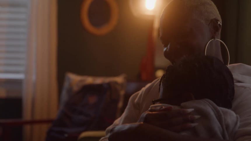 This is a mediumcloseup shot of a mother or older woman with short blonde hair and large hoop earrings cradling a child in a dimly lit cozy living room she is seen smiling down warmly at the child while a lamp provides a warm soft backligh