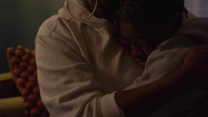 The video is a mediumcloseup shot of a darkskinned person in a lightcolored hoodie and glasses being embraced by another person showing a moment of intimate comfort the lighting is dim and moody with a textured orange pillow visible in the blurred