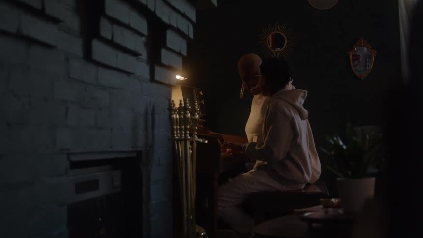 The video is a dimly lit medium shot of a person likely a musician in a lightcolored hoodie sitting on the right side of the frame and playing the piano or keyboard the light dramatically illuminates them against a dark brick wall on the left