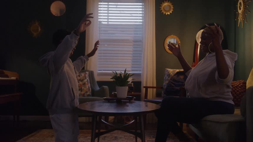 A woman sits on a couch opposite a teenage boy kneeling beside a small round table with a potted plant on it and both people raise their arms above their heads during an interaction in a dimly lit room