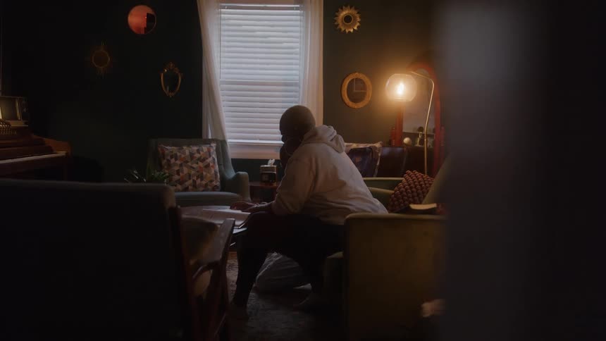A bald person in a white hoodie sits on a dark velvet couch helping a child writing with a pen in a notebook illuminated by a floor lamp in a dimly lit living room with dark green walls