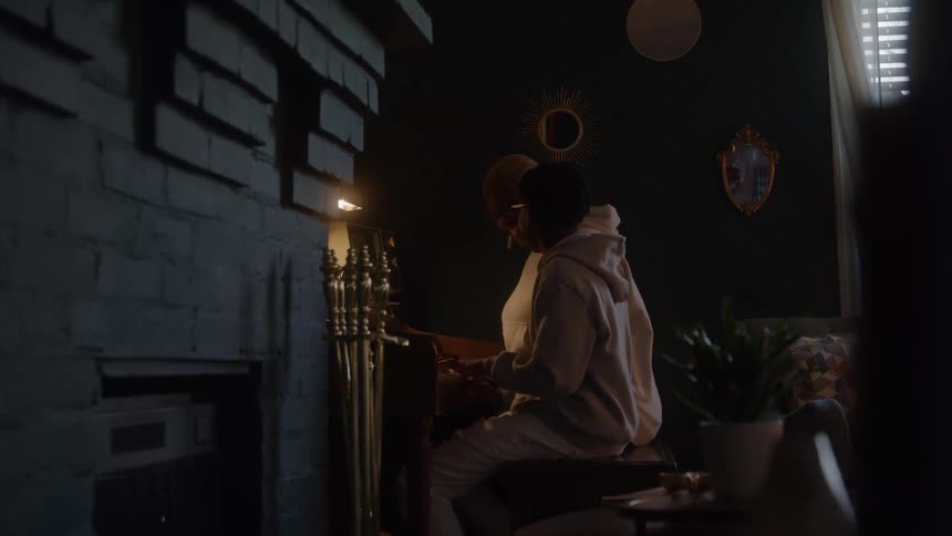 The video is a dimly lit medium shot of a person likely a musician in a lightcolored hoodie sitting on the right side of the frame and playing the piano or keyboard the light dramatically illuminates them against a dark brick wall on the left