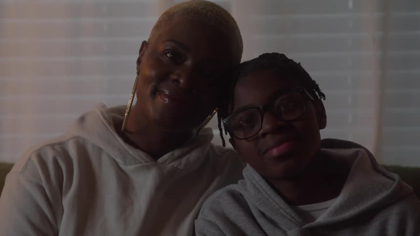 A happy woman with short blonde hair and large hoop earrings sits close to a boy wearing glasses and a gray hoodie with their faces illuminated softly against a dark background