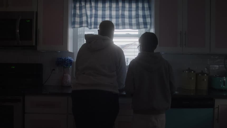 The video is a static medium shot from behind showing two people in hooded sweatshirts standing at a kitchen window the scene is dimly lit with bright daylight silhouetting them against the window which is covered by a blue and white plaid curtain