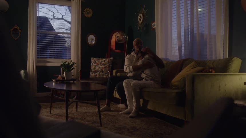 A woman and a teenage boy are sitting closely together on a long green couch with arms around each other in a dimly lit living room with two windows showing the dark outside