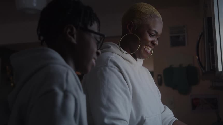 A happy woman with large hoop earrings and a boy wearing glasses in matching white and gray hoodies laugh while doing the dishes in a dimly lit indoor kitchen setting