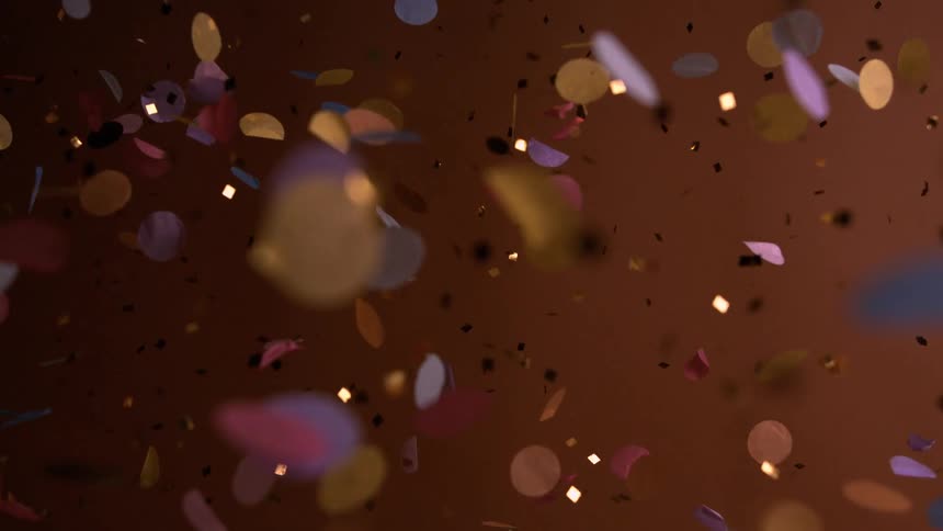 The video is a closeup shot of colorful confetti falling against a dark warm brown background the pieces are a mix of small circular and rectangular shapes in pink yellow blue and gold