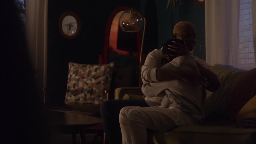 The video is a dimly lit mediumwide shot of a mother and son sitting close together on a sofa in a living room the mother has her arm around the son in a comforting gesture and she is seen affectionately wiping his face or eyes