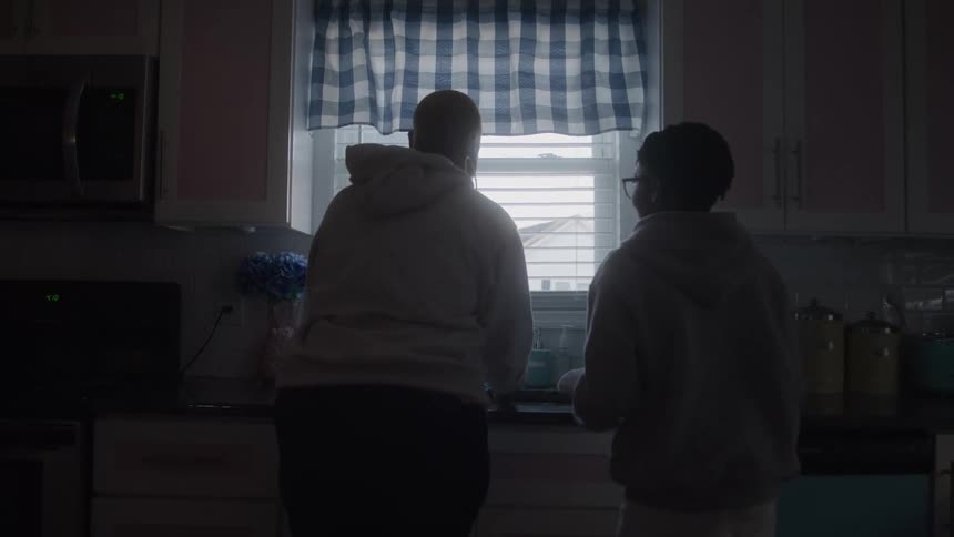 Two people stand side by side at a kitchen sink under a blue checkered valance backlit by the window as they work together