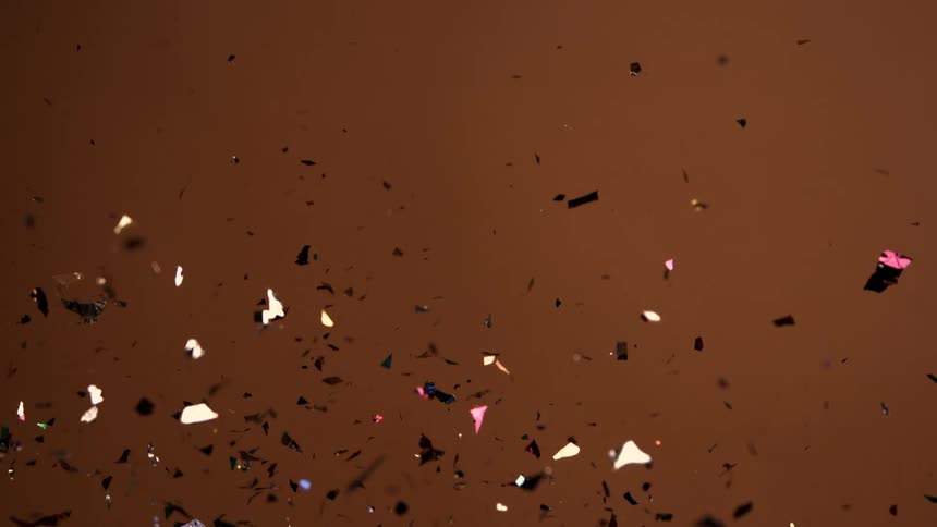 Numerous pieces of iridescent multicolored confetti with metallic and shiny textures fall gently downward against a warm brown background