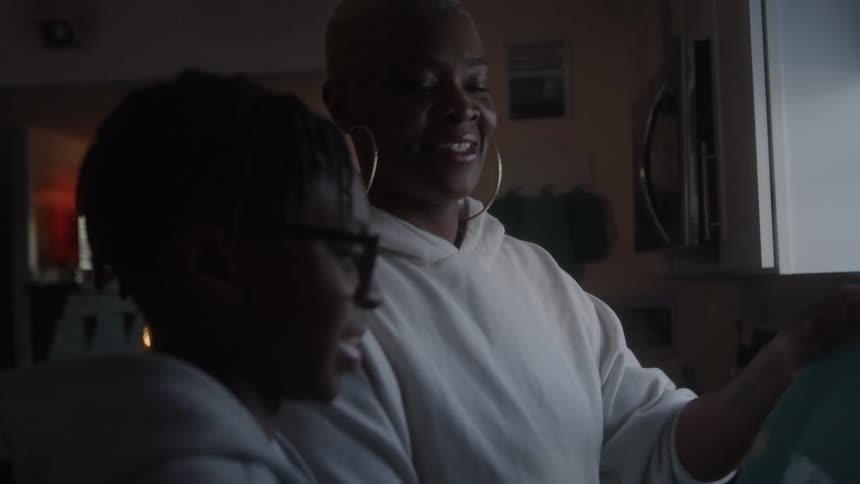 A woman with short light hair and large hoop earrings stands in a kitchen holding a light blue plate facing and smiling at a teenage boy wearing eyeglasses who is positioned in the foreground