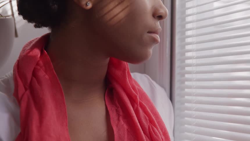 The video is a mediumcloseup profile shot of a young black woman with a low bun and a bright orangered scarf draped around her neck she is standing by a window with horizontal blinds and stripes of shadow fall across her face and shirt