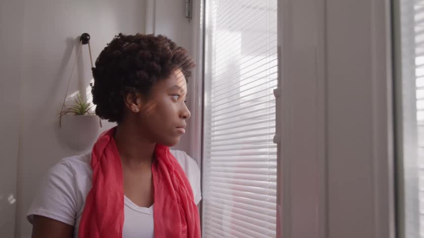 A woman in a red scarf stands by a window with blinds casting stripes on her face looking outside with a thoughtful slightly pensive expression