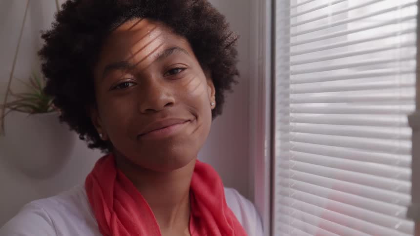A teenage girl with curly hair and a red scarf stands next to a window covered by horizontal blinds first looking to the right then turning the head and smiling