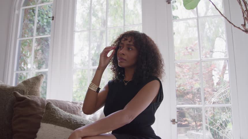 Woman with curly hair and golden bracelets sits near the bright window