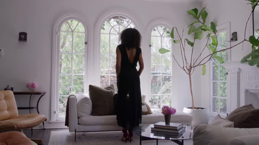 Woman standing in a sunny living room faces the windows while wearing a filmy black dress