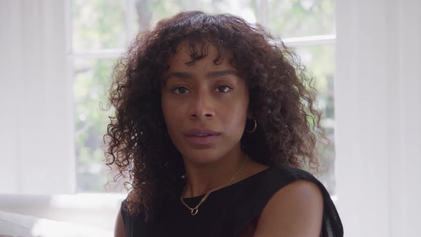 The video is a mediumcloseup of a young woman with curly hair opening her eyes and looking directly at the camera she is wearing a black sleeveless top and is positioned in front of a large bright window that provides natural light