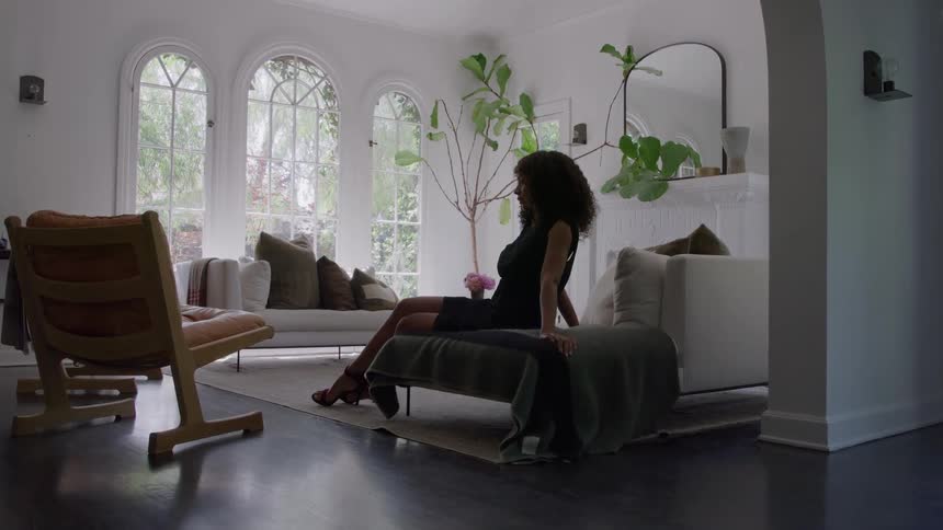 Woman wearing a black dress and red heeled shoes sits in a sunny living room