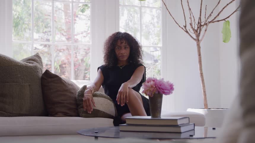 The video is a static medium shot of a young woman with curly hair sitting on a sofa in a brightly lit room with a large window she is dressed in a black top and jewelry looking offcamera with a contemplative expression with a vase of pink peonies vis