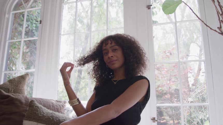 A woman with curly hair in a black sleeveless top sits near a couch in a bright windowed room looking relaxed and confident with a gentle smile