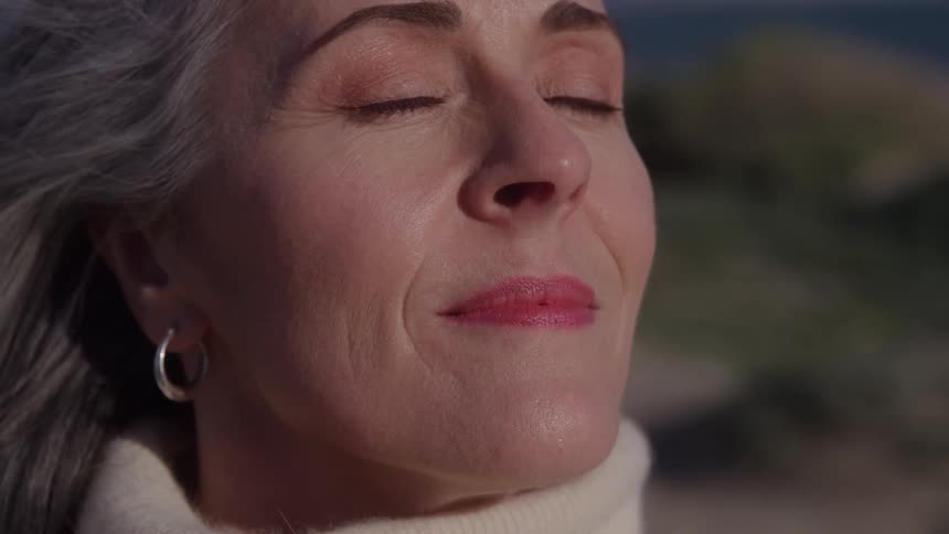 Closeup of a woman with silver hair and a hoop earring stands outdoors with eyes closed and a soft smile looking calm and content in a cream turtleneck