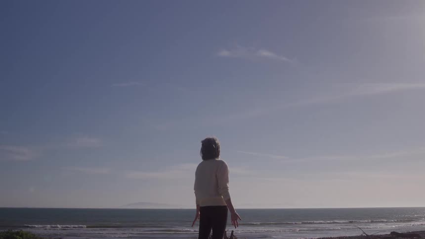 A woman stands on a beach back facing the camera as looks out at the vast ocean