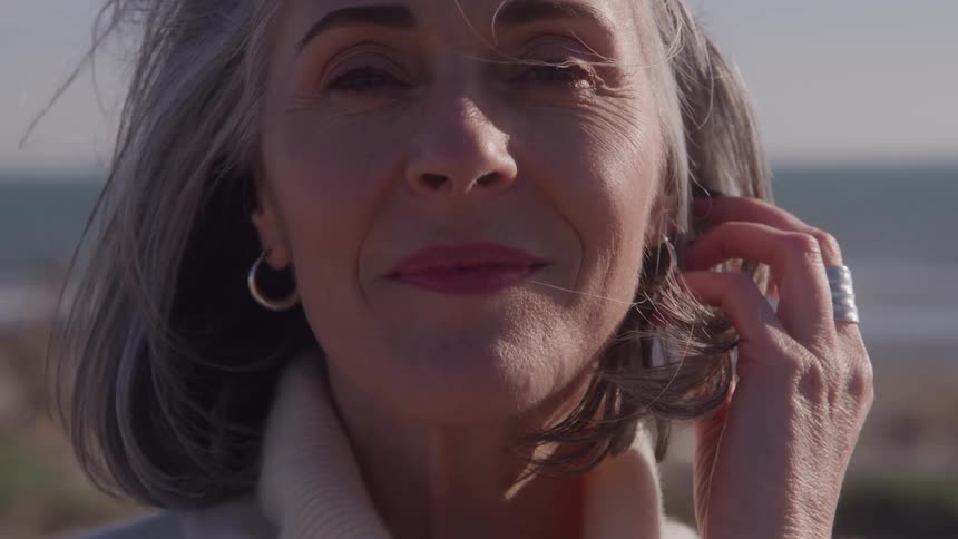 An elderly woman stands outdoors with a smile on her face gently stroking her grey hair