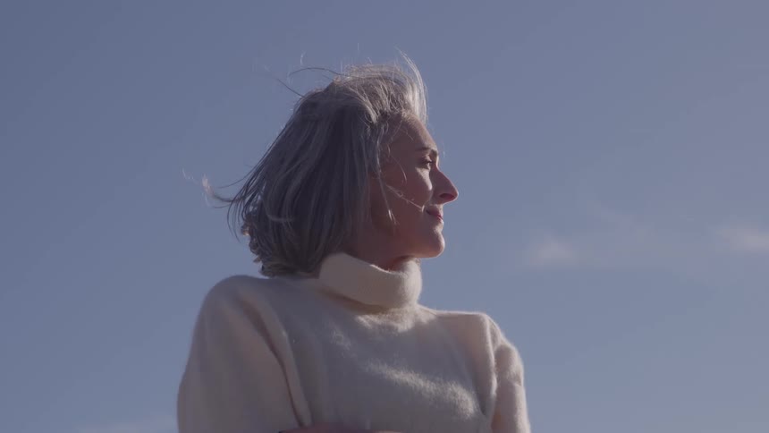 A mature woman wearing a white turtleneck sweater stands in a lowangle shot against a bright blue sky looking toward the right with arms folded while short gray hair is lifted by the wind