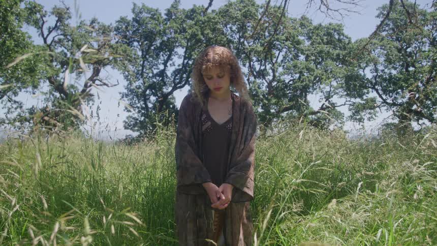A young woman stands alone in tall grass beneath oak trees with head bowed and hands clasped appearing sad and contemplative