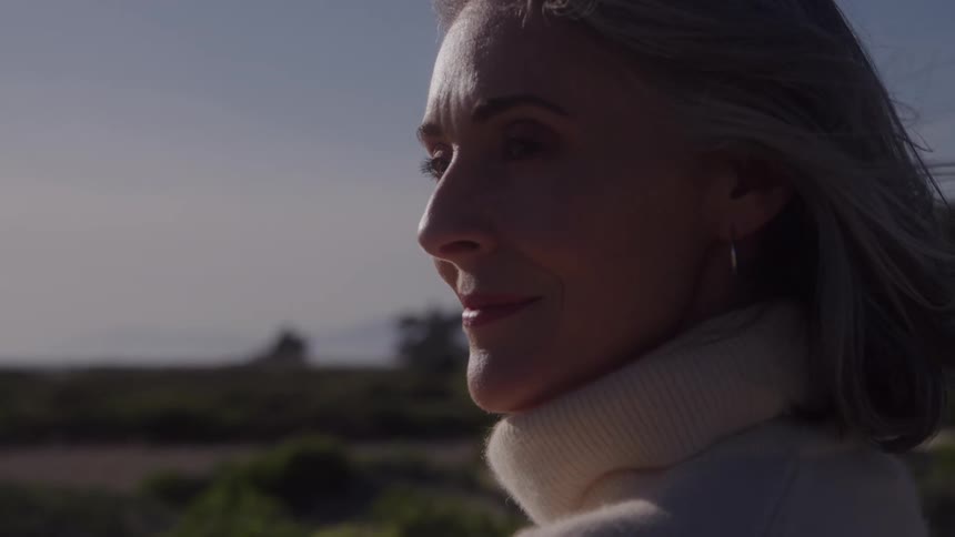 The video is a mediumcloseup profile shot of an older woman with gray hair wearing a white turtleneck standing outside on a sunny day her expression is peaceful and her hair is gently blowing in the breeze with a hazy blurred landscape in the back