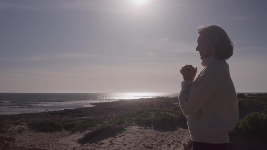 Woman gazes at the sea with hands clasped bathed in sunlight