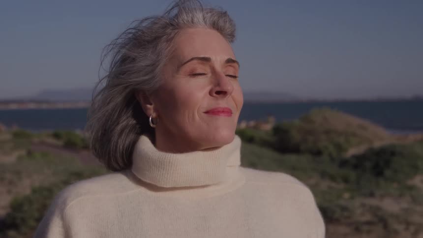 A woman with gray hair in a cream turtleneck stands by the coast with eyes closed and a faint smile looking peaceful in the sun