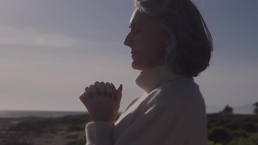 A woman with silver hair in a cream turtleneck stands in profile outdoors hands clasped near her chin and eyes closed wearing a serene grateful expression in soft evening light