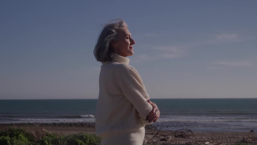 The video is a mediumwide profile shot of an older woman with gray hair standing outdoors against a coastal landscape she is wearing a lightcolored turtleneck sweater and has her arms crossed looking out at the ocean on a slightly breezy day