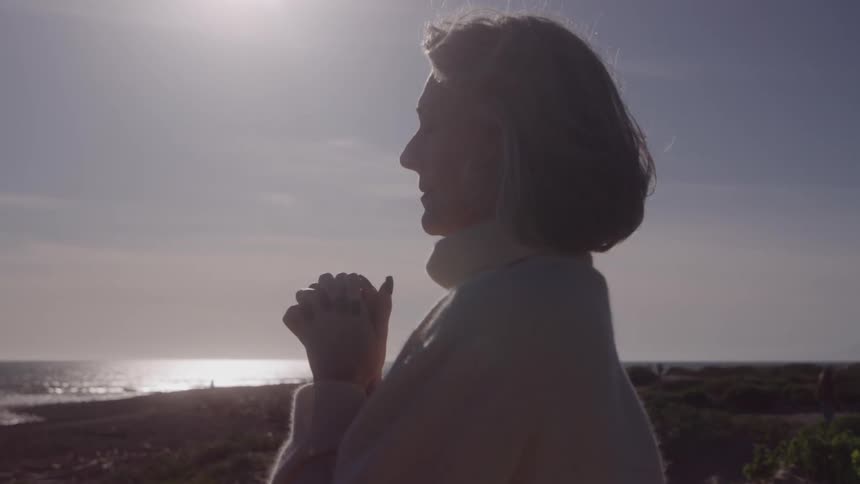 The video is a mediumcloseup profile shot of an older woman standing outdoors with the ocean in the background she is silhouetted against a bright sun over the water and her hands are clasped near her face in a contemplative or prayerful gesture