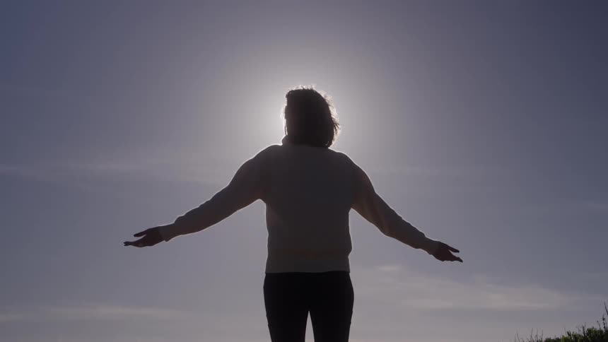 Silhouette of a person stands with arms outstretched against the sun outdoors