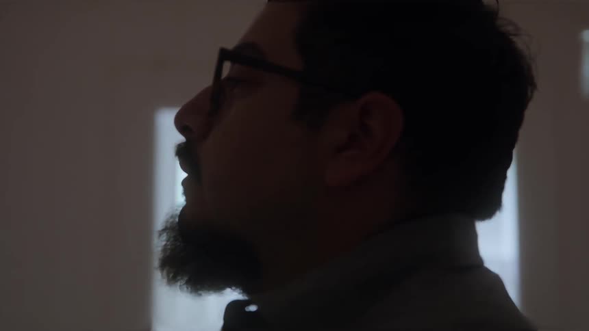 The video is a mediumcloseup profile shot of a mans face in a dimly lit room he is wearing glasses and has a prominent beard with a sliver of light illuminating the side of his face against the darkness