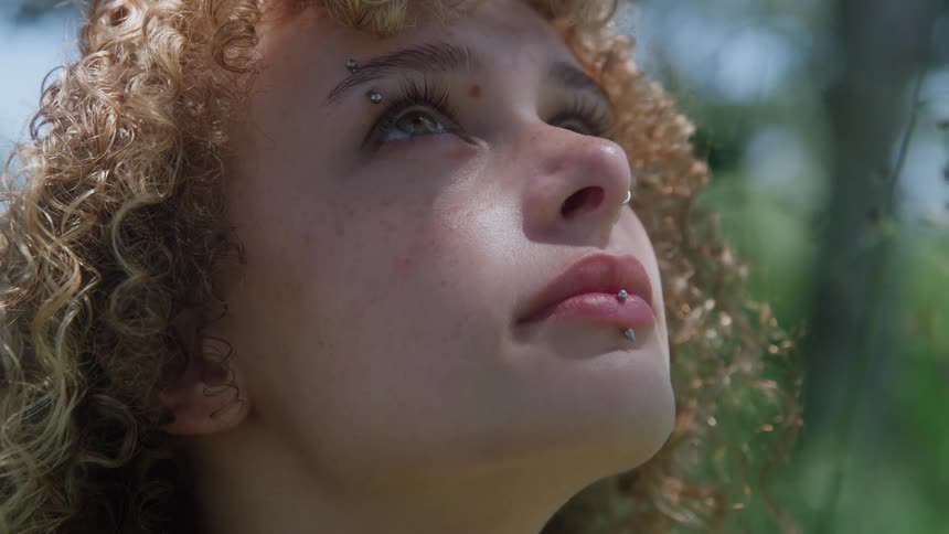 Closeup of a young woman with curly hair and facial piercings looking upward appearing thoughtful and hopeful