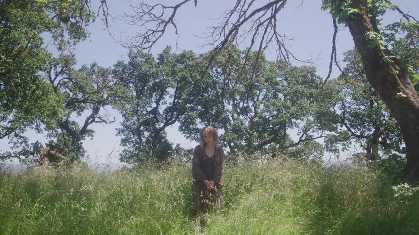 Woman stands passively in tall grass and trees under a bright blue sky