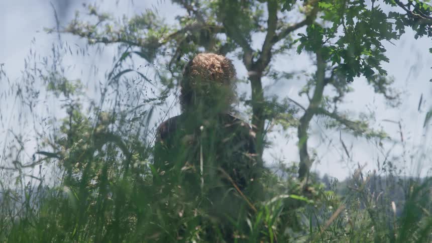 The video is a lowangle mediumcloseup shot of a person standing outdoors in a grassy area the person who has curly reddish hair is partially obscured by tall blades of grass and leaves with trees visible in the background against a bright sky
